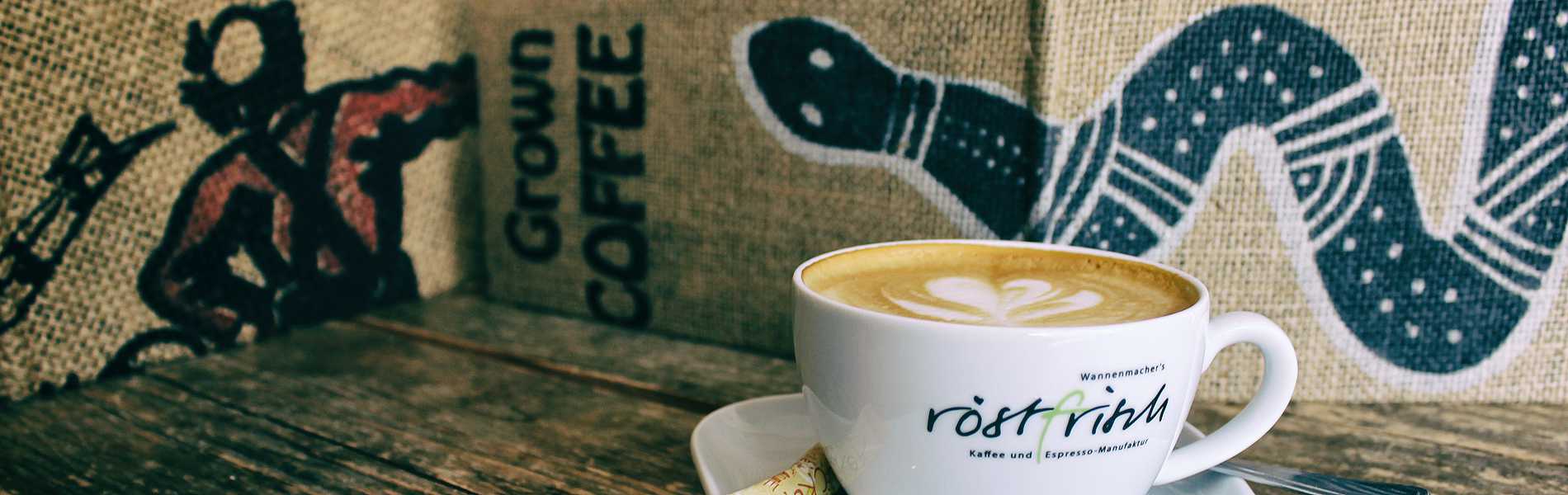 Cappuccino mit Latte Art in einer röstfrisch-Tasse vor Jutesäcken mit Rohkaffee